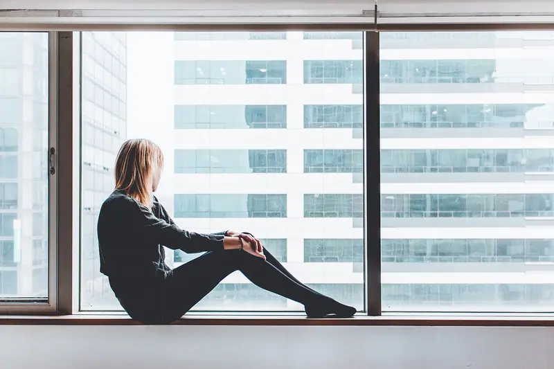 Pessoa sentada no parapeito de uma janela ampla, de lado, olhando os prédios da cidade num dia claro