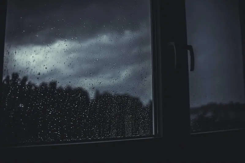 Canto de uma janela escura com gotas de chuva no vidro e céu nublado ao fundo