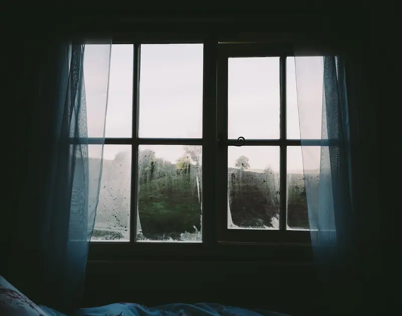 Janela de quarto vista de dentro, com cortinas claras e os cantos do vidro embaçados