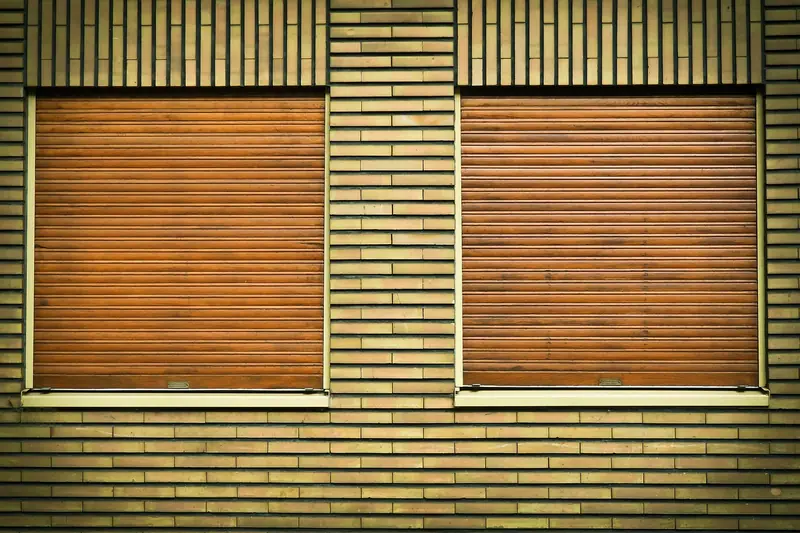 Duas janelas com persianas de enrolar de cor marrom fechadas numa parede de tijolos