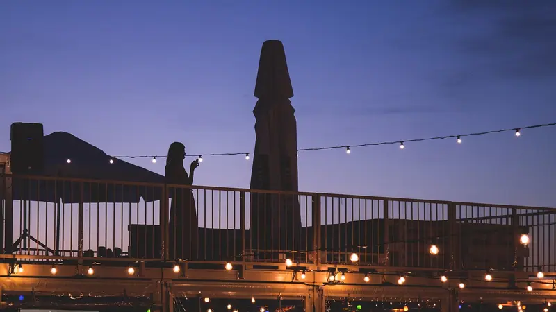 Terraço de bar ao entardecer, com cordão de lâmpadas acesas e pessoas ao longe