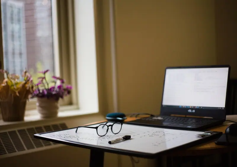 Notebook aberto sobre a mesa, ao lado de uma janela com vasos de flores no parapeito