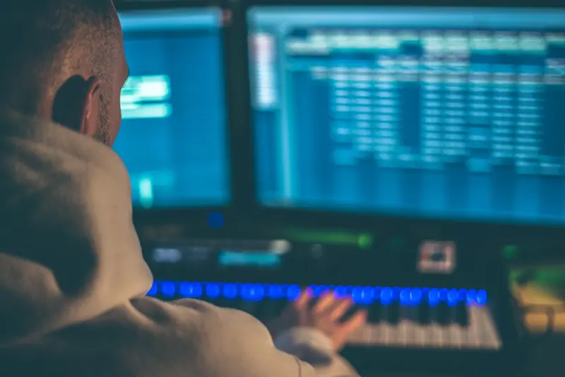 Pessoa de costas diante de monitores com programa de áudio e um teclado musical iluminado