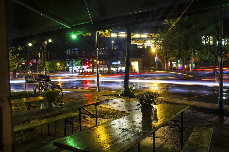 Mesas e bancos de madeira na calçada de um bar à noite, com a rua molhada ao fundo