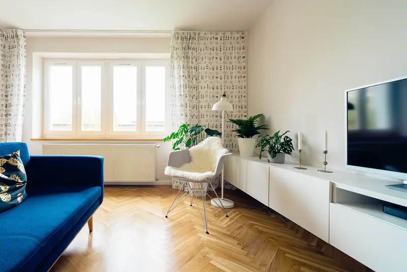 Sala com sofá azul, piso de taco e janela clara com cortina leve, plantas e rack branco