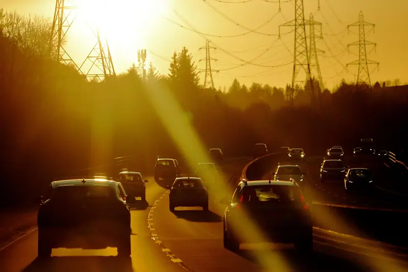 Carros enfileirados numa avenida sob a luz forte do fim de tarde, com torres de energia ao fundo