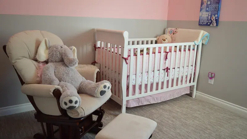 Quarto de bebê com berço branco, poltrona de amamentação com bichos de pelúcia e paredes em tom rosa claro