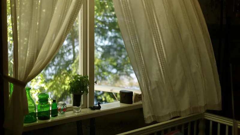 Cortinas claras balançando com o vento diante de uma janela, com vasos de plantas no parapeito