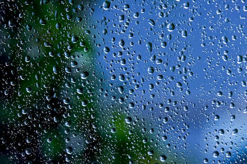 Gotas de chuva espalhadas por um vidro de janela, com o céu azul e folhas verdes desfocados atrás