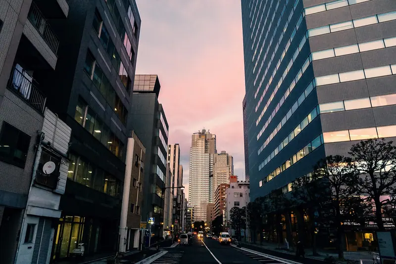 Rua entre prédios altos de escritórios e apartamentos ao entardecer, com o céu rosado ao fundo