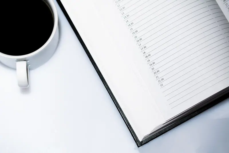 Agenda aberta com os horários do dia em branco, ao lado de uma xícara de café preto, sobre uma mesa branca