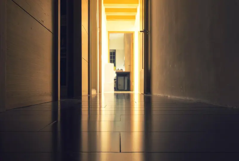 Corredor estreito e escuro, com piso brilhante e uma porta aberta iluminada no fim