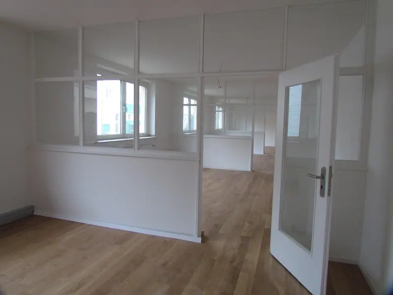 Sala de escritório vazia com piso de madeira, divisórias brancas e uma porta branca aberta