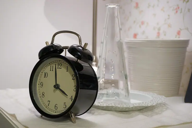 Despertador preto marcando quatro horas sobre o criado-mudo, ao lado de um vaso de vidro