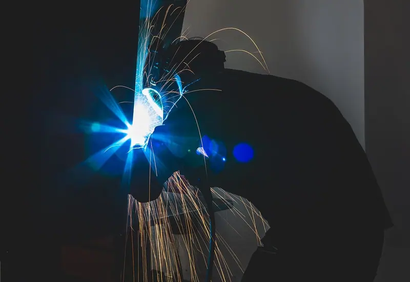 Silhueta de uma pessoa soldando metal, com faíscas e um clarão azul no escuro