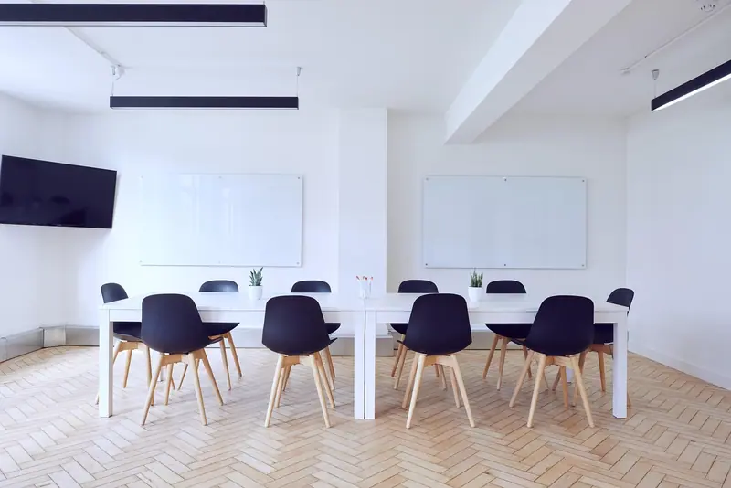 Sala de reunião clara com mesa comprida, cadeiras pretas e televisão na parede