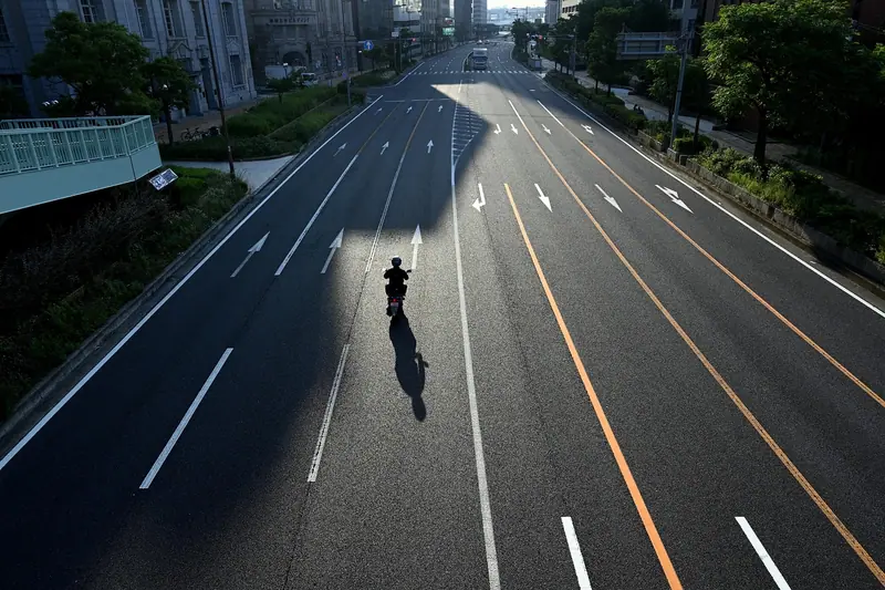 Avenida larga quase vazia vista do alto, com uma única moto e a sombra dos prédios no asfalto