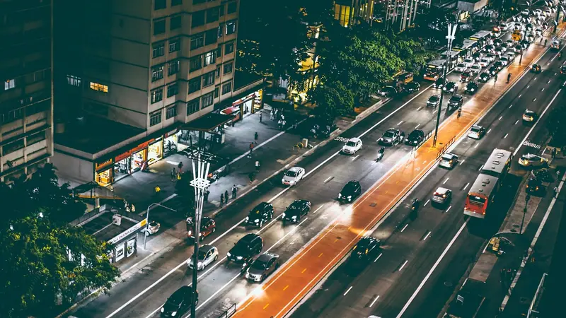 Avenida larga com muitos carros vista do alto, entre prédios e árvores, à noite