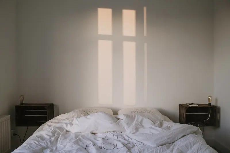 Cama arrumada com edredom branco e a luz de uma janela desenhada na parede do quarto