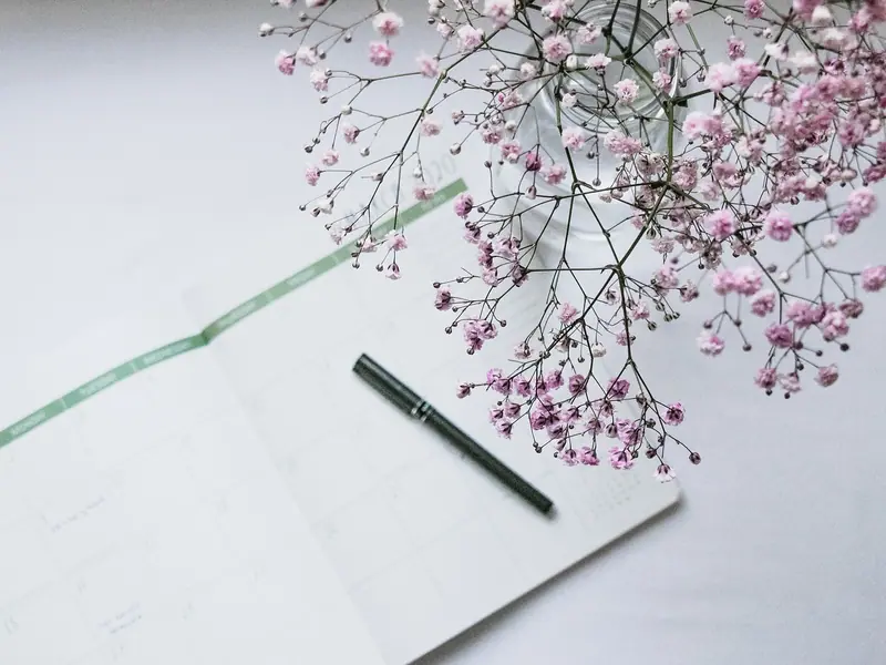 Agenda mensal aberta com uma caneta preta e um vaso de flores miúdas ao lado