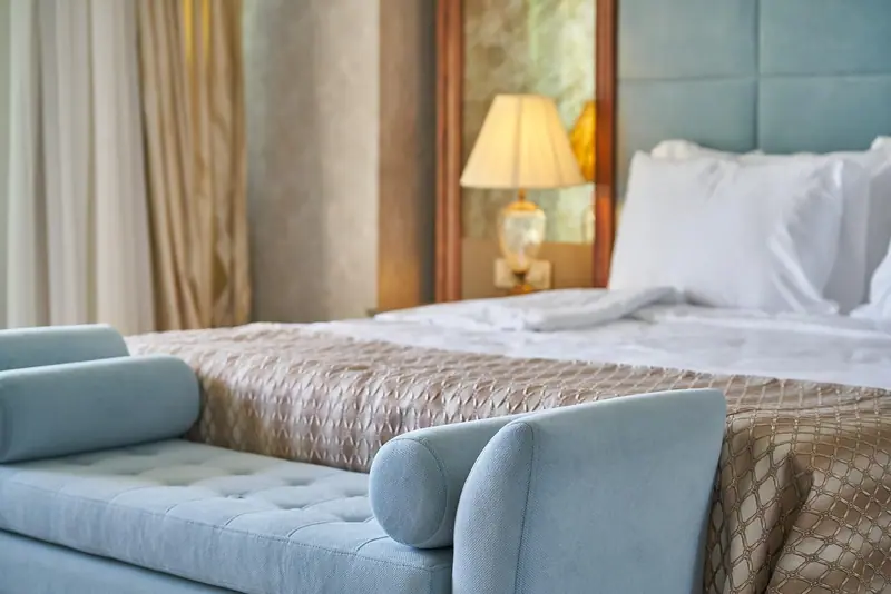 Cama de casal com travesseiros brancos, abajur aceso ao lado e um banco estofado azul ao pé da cama