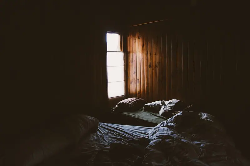 Quarto escuro com cama desarrumada e uma janela pequena deixando entrar a luz do dia