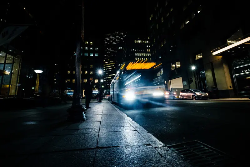 Ônibus iluminado passando em movimento por uma rua escura, de madrugada, com prédios ao fundo