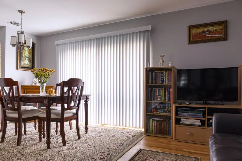 Sala de estar com mesa de jantar de madeira, estante, televisão e persiana vertical na janela