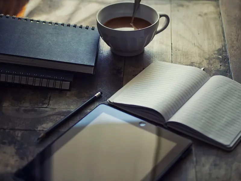 Caderno aberto, tablet e xícara de café sobre uma mesa de madeira escura