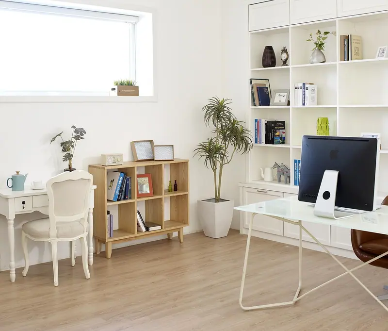 Escritório em casa claro, com mesa branca, computador, estante com livros e plantas junto à janela