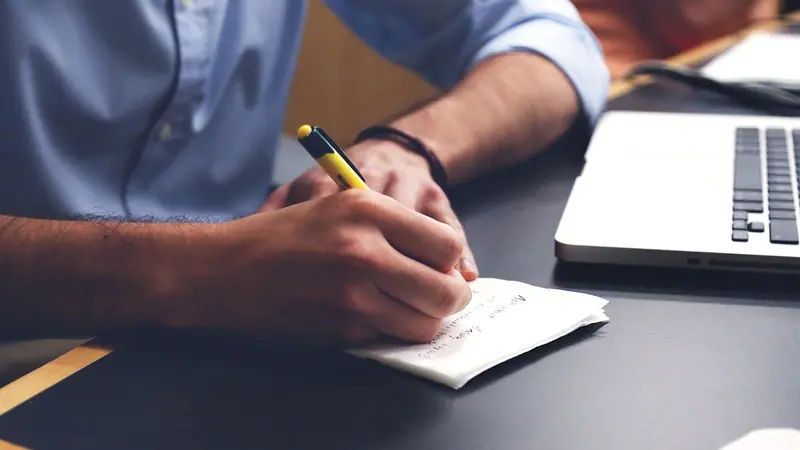Mão segurando uma caneta e anotando num papel, ao lado de um notebook sobre a mesa