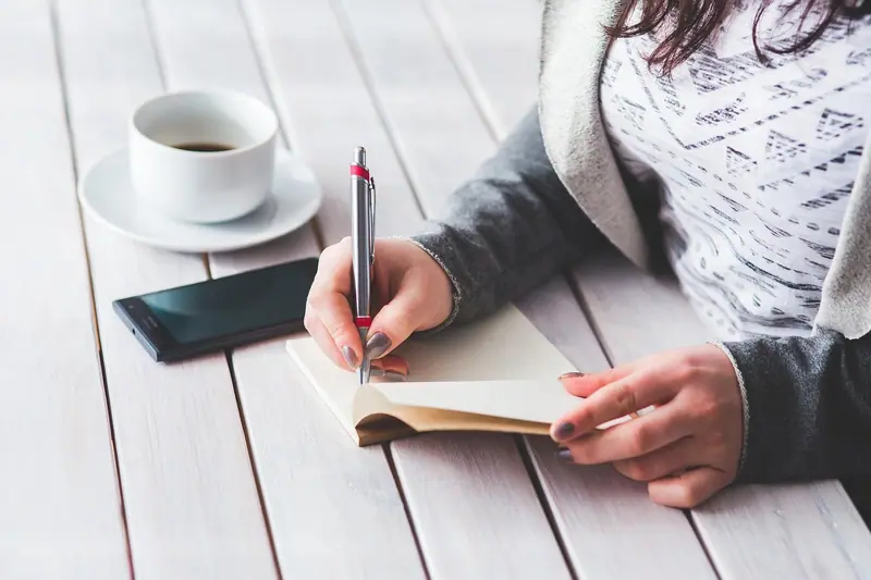 Mão de mulher escrevendo num caderno pequeno, ao lado de uma xícara de café e de um celular