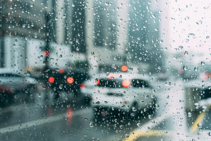 Carros numa rua movimentada vistos através de um vidro com gotas de chuva