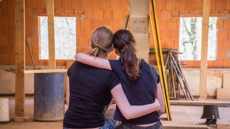 Duas pessoas sentadas de costas, abraçadas, olhando um cômodo em obra com paredes de tijolo à mostra