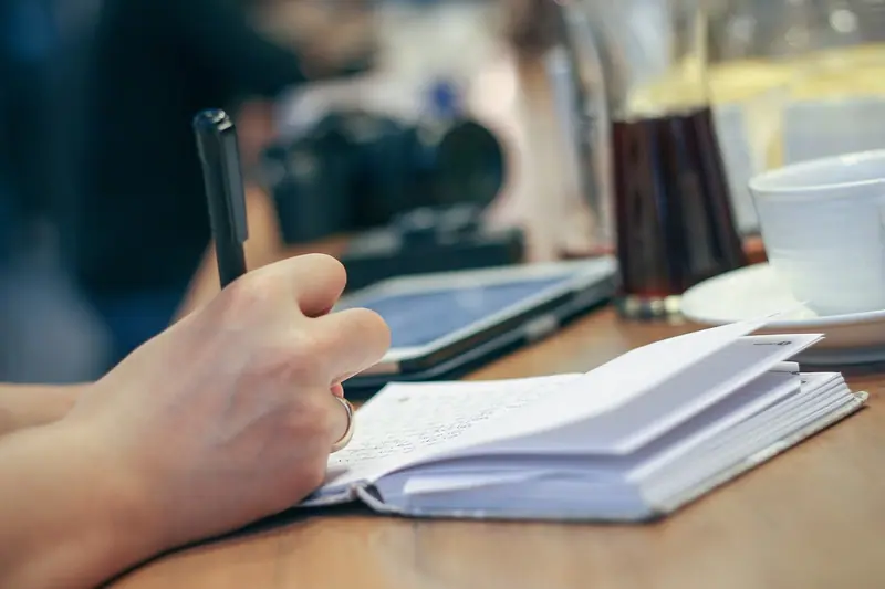 Mão escrevendo com caneta num caderno aberto sobre uma mesa de madeira
