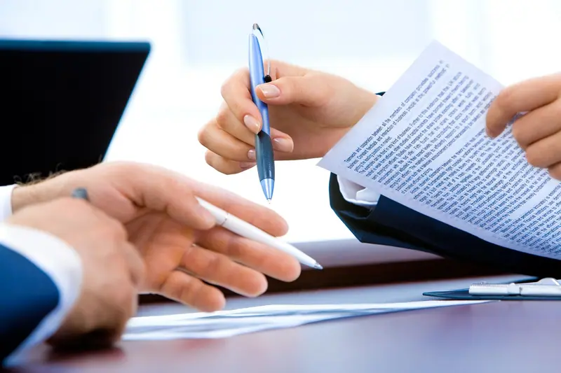 Mãos segurando uma caneta e folhas impressas sobre uma mesa de escritório