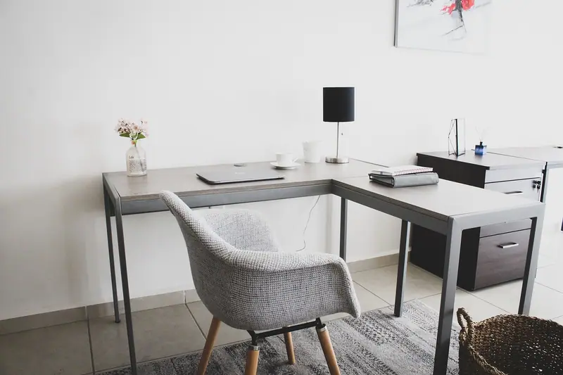 Sala clara com mesa de trabalho em L, cadeira estofada cinza e luminária preta