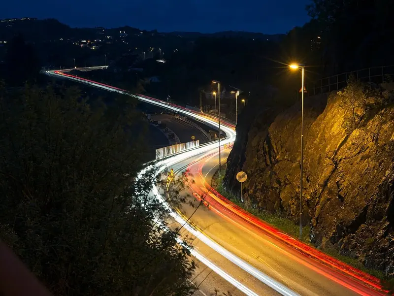 Estrada em curva à noite, com rastros de luz dos faróis entre árvores e um paredão de pedra