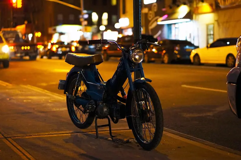Moto pequena estacionada na beira da calçada à noite, com faróis de carros e luzes da cidade desfocados ao fundo