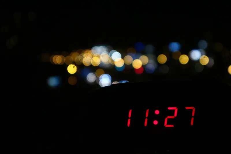 Relógio digital com números vermelhos num cômodo escuro, com luzes da cidade desfocadas atrás