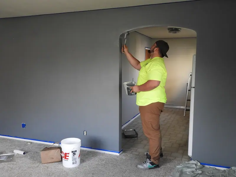 Pintor de costas pintando uma parede cinza de um apartamento em reforma, com balde e rolo no chão