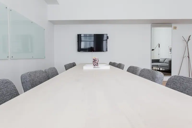 Sala de reunião com mesa comprida, cadeiras cinza, televisão na parede e divisória de vidro à esquerda