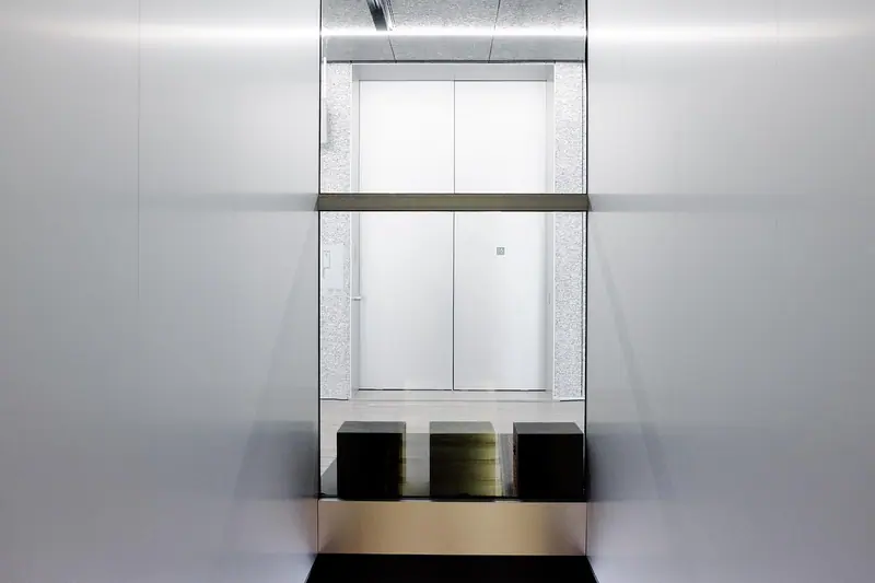 Hall de elevador com portas metálicas fechadas, paredes claras e três bancos escuros