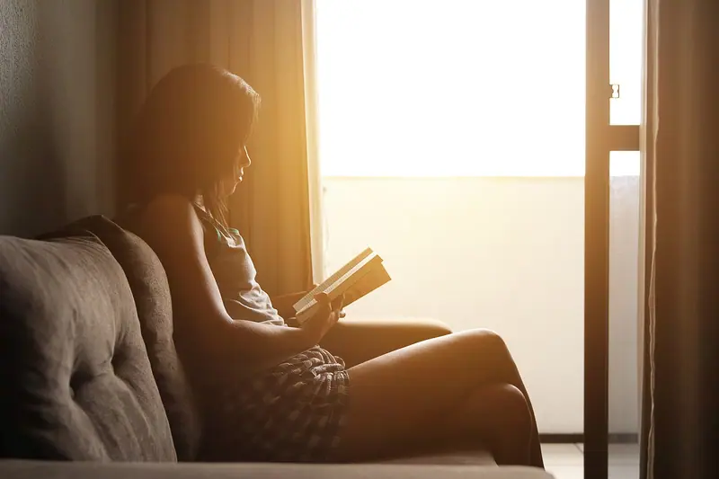Silhueta de uma pessoa lendo um livro no sofá, ao lado de uma janela iluminada