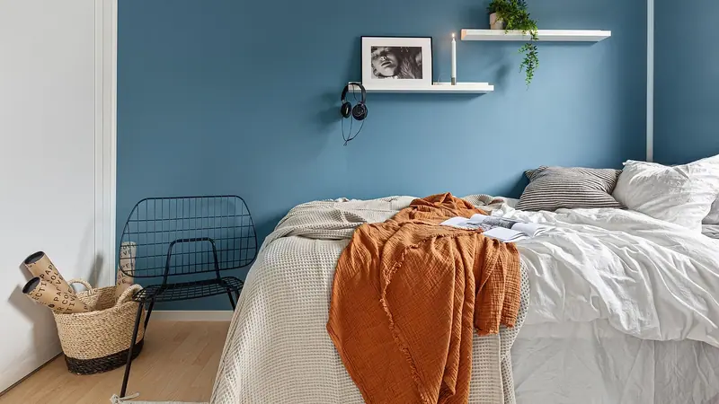 Quarto tranquilo com parede azul, cama de lençóis brancos e manta laranja, iluminado pela luz do dia