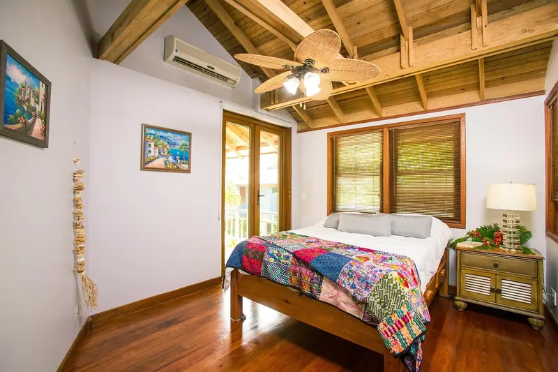 Quarto com cama de colcha colorida, ventilador de teto e ar-condicionado no alto da parede