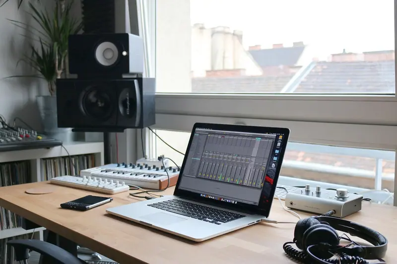 Home studio com notebook, caixas de som e fones sobre uma mesa de madeira, ao lado de uma janela