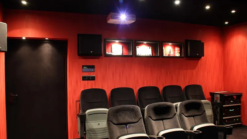 Sala de home theater com fileiras de poltronas de cinema, paredes vermelhas, caixas de som e porta escura ao lado