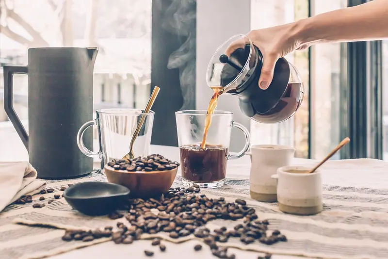 Mão servindo café numa xícara sobre a mesa, com grãos e bule ao redor