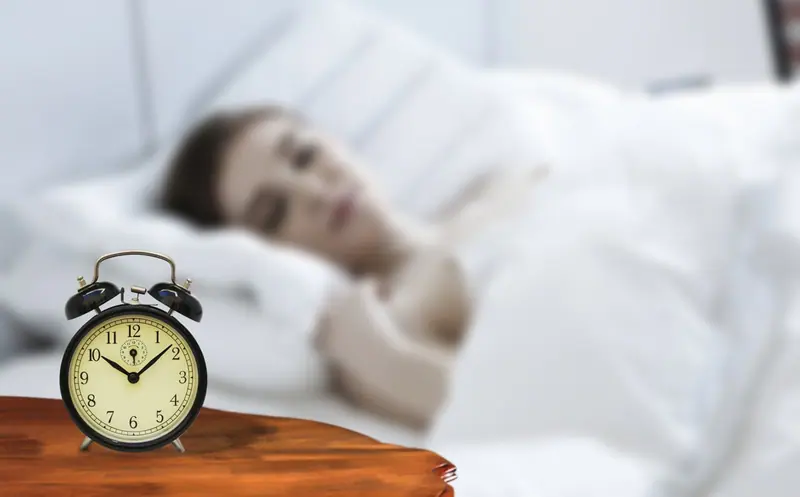 Despertador antigo ao lado da cama, com uma pessoa dormindo desfocada ao fundo